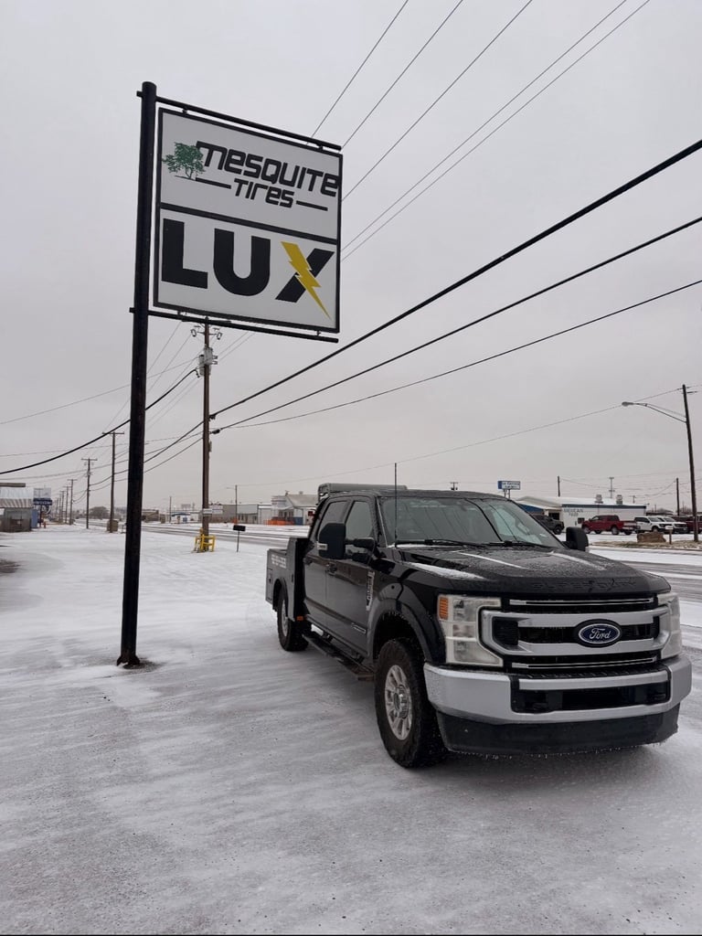 Black and white Ford pickup truck parked under a Mesquite Lux dealership sign on a rainy day
